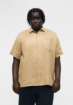 Muž s tmavou pokožkou a krátkymi dredmi, oblečený v béžovej košeli s krátkym rukávom a tmavých nohaviciach, stojaci pred jednoduchým pozadím.