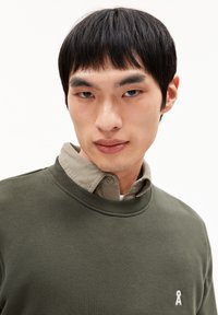Olijfgroene sweatshirt met ronde hals, voorzien van een klein wit logo aan de linkerkant. Draag je over een lichtbeige overhemd met kraag.