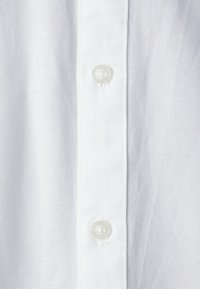 Camicia bianca con tessuto testurizzato. Presenta una fila di bottoni trasparenti e rotondi e un design pulito e semplice.