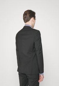 Giacca nera con un design aderente, tessuto liscio e rever a punta. La camicia sotto presenta un motivo a quadretti.