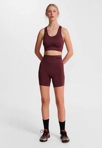 Bordeauxrode sportbeha en bijpassende korte broek met tailleband, met een gladde textuur en subtiele branding. Draagt zwarte enkelsokken en sportschoenen.