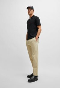 Black polo shirt with a zipper detail, beige straight-leg pants, and black loafers. Model wears a black cap with front logo.