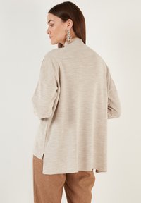 Cardigan tricotat bej, cu guler înalt, croi lejer și umeri căzuți. Dispune de fante laterale pentru un plus de libertate de mișcare și confort.