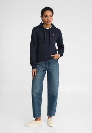 Femme portant un sweat à capuche bleu marine, un jean droit bleu et des baskets beiges, debout avec une main dans sa poche.