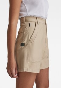 Béžové plátěné šortky s přiléhavým pasem, bočními kapsami a viditelným šitím; látka má hladkou strukturu a čistý povrch.