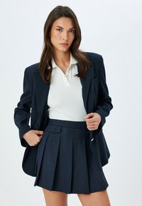 Marineblazer met zijzakken over een witte polo, gecombineerd met een geplooide marine rok met een knoop en textuurstof.