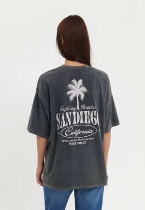 Personne aux longs cheveux bruns, vue de dos, portant un T-shirt oversize gris foncé avec une inscription blanche « San Diego California » et un motif de palmier, ainsi qu’un jean bleu.