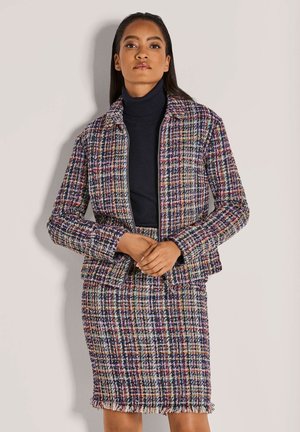 Vrouw draagt een meerkleurig geruit jack en bijpassende rok over een zwarte coltrui, staat met gevouwen handen voor zich.