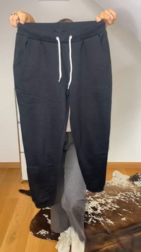 Persona sosteniendo pantalones de chándal negros con cordones blancos, de pie sobre un suelo de madera cerca de una alfombra de piel de vaca.