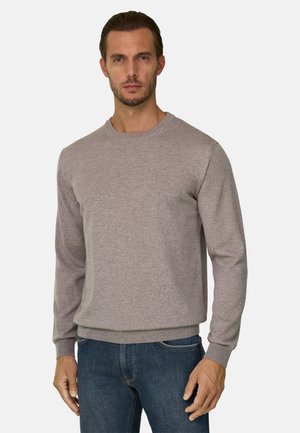 Homme portant un pull col rond marron clair et un jean bleu, debout devant un fond clair uni.
