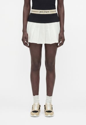 Osoba nosząca czarny top z białym paskiem z napisem „Palm Angels”, białą plisowaną spódnicę, białe skarpetki oraz beżowo-czarne trampki.