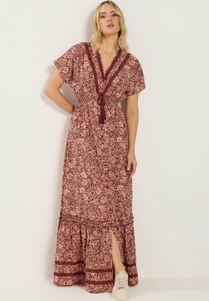 Donna che indossa un abito lungo floreale color melanzana e beige con maniche corte, scollo a V, laccio con nappine, spacco e scarpe da ginnastica bianche davanti a uno sfondo neutro.