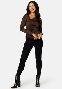 Top marrone a maniche lunghe con design arricciato, scollo a V e dettaglio con lacci. Abbinato a jeans skinny neri e stivaletti alla caviglia.