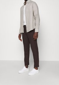 Camisa de lino gris claro abotonada sobre una camiseta blanca, combinada con pantalones oscuros marrones y zapatillas blancas. Bolsillo visible en la camisa.