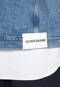 Džínová bunda s světle modrým omakem, bílou etiketou s nápisem "CALVIN KLEIN JEANS" a kontrastním prošíváním podél švů.
