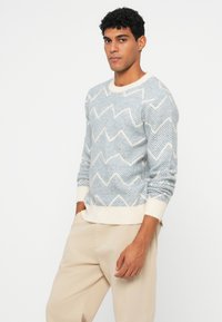 Jasnoniebieski sweter z wzorem w zygzak, w różnych odcieniach, z kremowymi ściągaczami i dołem, wykonany z materiału o teksturowanym splocie.