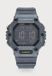 Montre numérique avec un écran rectangulaire noir et un bracelet en caoutchouc gris. Les caractéristiques incluent une résistance à l'eau, des fonctions d'alarme et une autonomie de 10 ans.