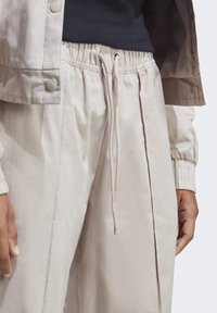 Ljust beige, lätta byxor med elastisk midja, dragsko, sidofållar och en slät, något blank yta.