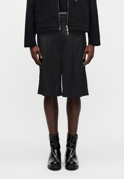 Μαύρο κοντό μπουφάν με κλείσιμο με φερμουάρ, σετ με πλισέ μαύρα σορτς και γυαλιστερές μαύρες μποτάκια αστραγάλου. Απλός, μινιμαλιστικός σχεδιασμός.