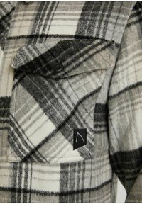 Ruudullinen flanellipaita harmaana ja kermaisen valkoisena, keskipisteenä rintatasku mustilla painikkeilla, pehmeä tekstuuri ja logo-patch etupuolella.