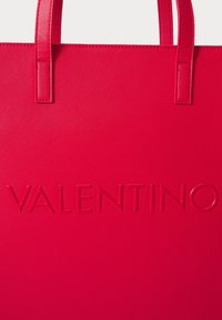 Röd tote-väska med strukturerad yta, med det präglande "VALENTINO"-logotypen på framsidan och två robusta handtag.