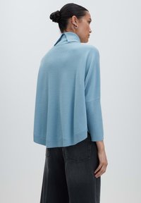 Frau mit einem Dutt, die einen hellblauen, lockeren Pullover mit hohem Kragen und schwarzen, weiten Jeans trägt, seitlich blickend.