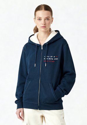 Sweat à zip bleu marine avec une doublure en polaire blanche douce, deux poches avant et un texte imprimé en rouge et blanc sur la poitrine.