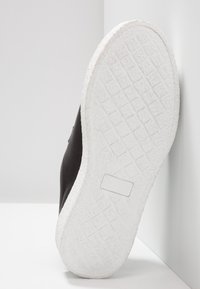 Svart sneaker med en texturerad, vit gummisula, som har en låg profil och en slät läderöverdel. Sulan har ett rutmönster för bra grepp.