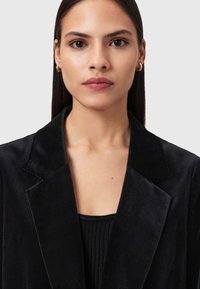 Giovane donna con capelli neri lisci che indossa orecchini a cerchio d'oro, un blazer di velluto nero e un top nero a coste, rivolta in avanti con un'espressione neutra.
