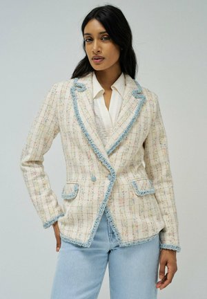 Blazer en tweed couleur crème avec des accents de fil colorés, un bord effiloché bleu, des revers à cran et deux poches avant, porté sur une chemise blanche.