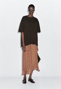 Kobieta w oversize'owej czarnej bluzie z krótkim rękawem, brązowej kratkowanej spódnicy midi, czarnych loafersach, w okularach przeciwsłonecznych, trzymająca ciemną torebkę na tle jednolitym.