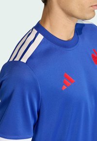 Modré sportovní tričko z hladké látky. Má bílé třípruhové akcenty na ramenech a červené detaily s logem, se kulatým výstřihem.