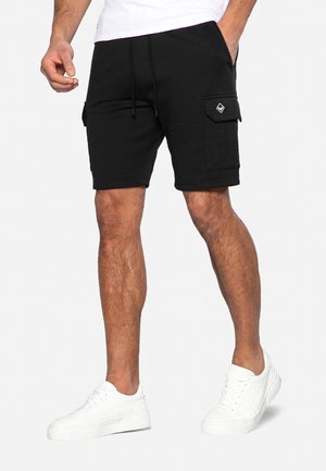 Shorts cargo noirs en mélange de coton, avec des poches latérales, une taille élastique avec cordon de serrage, et une texture lisse. Associés à des baskets blanches.
