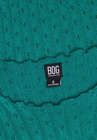 Tyrkysový pletený svetr s ribbovanou texturou a malými otvory. Má rýhovaný výstřih a štítek s textem "BDG, Urban Outfitters, S, vyrobeno v Turecku."
