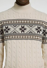 Krämfärgad tubhalsad tröja med kabelstickad struktur, som har ett mörkbrunt och svart geometriskt mönster över bröstet.
