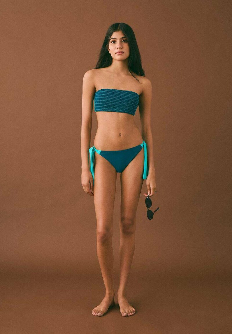 Bikini set vključuje strukturirano modro bandeau zgornji del in temno modre hlačke z svetlomodrimi bočnimi vrvicami; model drži črne sončna očala, noge so bose.