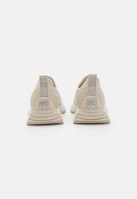 Sportssko med en beige syntetisk overdel, teksturert hælflik og en kontrasterende hvit gummisåle med et DKNY-logo på baksiden.