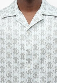 Κοντινή λήψη ενός ανοιχτό μπλε πουκάμισου με κουμπιά, με επαναλαμβανόμενο σχέδιο από ανοιχτό γκρι διαπλεκόμενα γράμματα και φαρδιά γιακά.