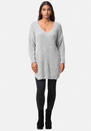 Kvinde iført en lysegrå oversized striktrøje, sorte skinny bukser og sorte ankelstøvler med hæl mod en ensfarvet baggrund.