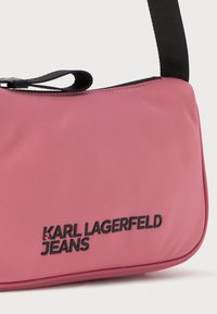 Rozā neilona plecu soma ar melnu siksnu. Uz priekšpuses ir melni izsisti uzraksti "KARL LAGERFELD JEANS" un gluda, matēta tekstūra.