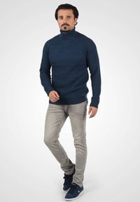 Tumši zila adīta augsto apkakliņu džemperis ar teksturētu rakstu, kombinēts ar gaiši pelēkām šaurām džinsām un tumši ziliem sporta apaviem.