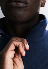 Persoon die een blauwe fleecejas draagt, hand trekt de rits omhoog naar de nek, lippen en kin zichtbaar, neutrale achtergrond.