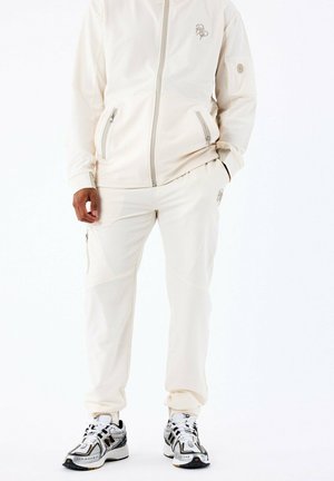 Chaqueta de cremalleras en color crema claro con dos bolsillos frontales y un logo bordado, combinada con joggers a juego que presentan costuras contrastantes y un bolsillo con cremallera.