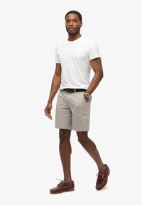 Hvid T-shirt med rund hals, beige shorts, sort bælte og burgunder bådsko. En bemærkelsesværdig armbåndsur tilføjer farve. Modellen står foran en neutral baggrund.