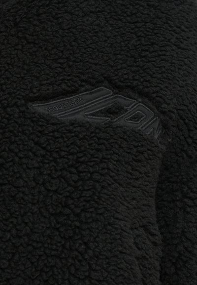 Veste en polaire noire avec une surface texturée et un patch logo visible. Le tissu semble doux et épais, ce qui améliore chaleur et confort.