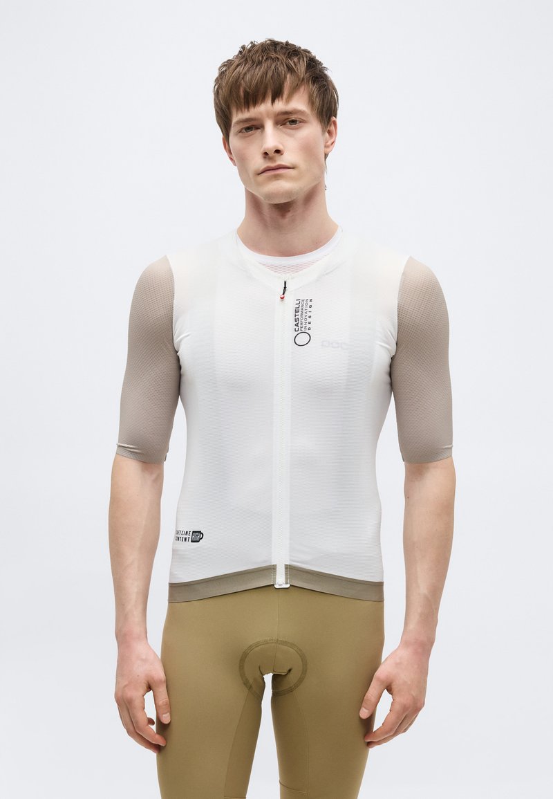 Giovane uomo che indossa una maglia da ciclismo bianca a maniche corte e pantaloncini da ciclismo imbottiti beige, in piedi davanti a uno sfondo chiaro e uniforme.