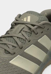 Scarpa sportiva marrone chiaro con tomaia in mesh, lacci incrociati e tre strisce color crema sul lato, mostrata in primo piano.