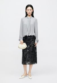 Hellgraue Bluse mit Schluppe, schwarzer Bleistiftrock mit Pailletten, weißes Clutch-Täschchen und beigefarbene spitze Pumps, vor schlichtem Hintergrund.