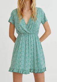 Robe florale verte à manches courtes, décolleté en v, taille élastique et ourlet à volants. Présente de petits motifs floraux blancs et roses partout.