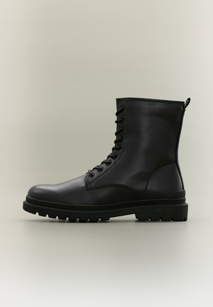 Bottine noire en cuir à lacets, dotée d’une semelle épaisse à crampons, présentée de profil sur un fond clair uni.